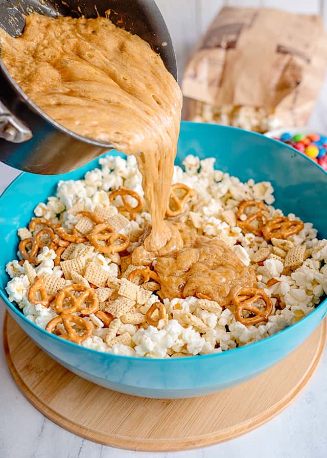 homemade caramel being poured into blue bowl with popcorn, cereal, and pretzels