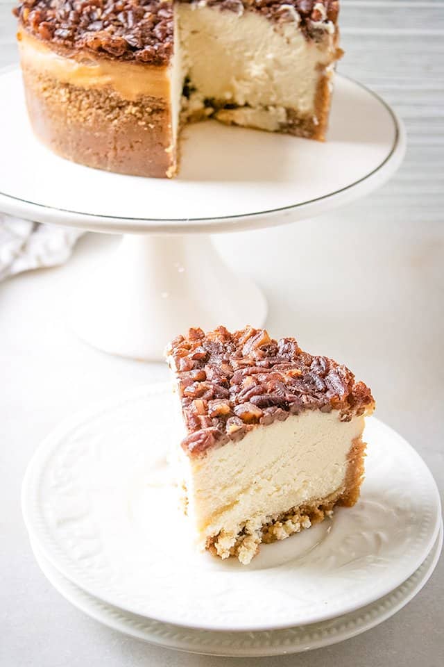 pecan pie cheesecake sitting on large white plates with a cake plate behind it with the remaining cheesecake on it