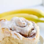 closeup of bananas foster cinnamon rolls on a white plate with frosting and a banana sitting next to it
