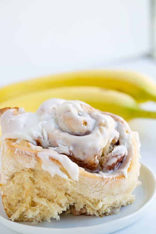 closeup of bananas foster cinnamon rolls on a white plate with frosting and a banana sitting next to it