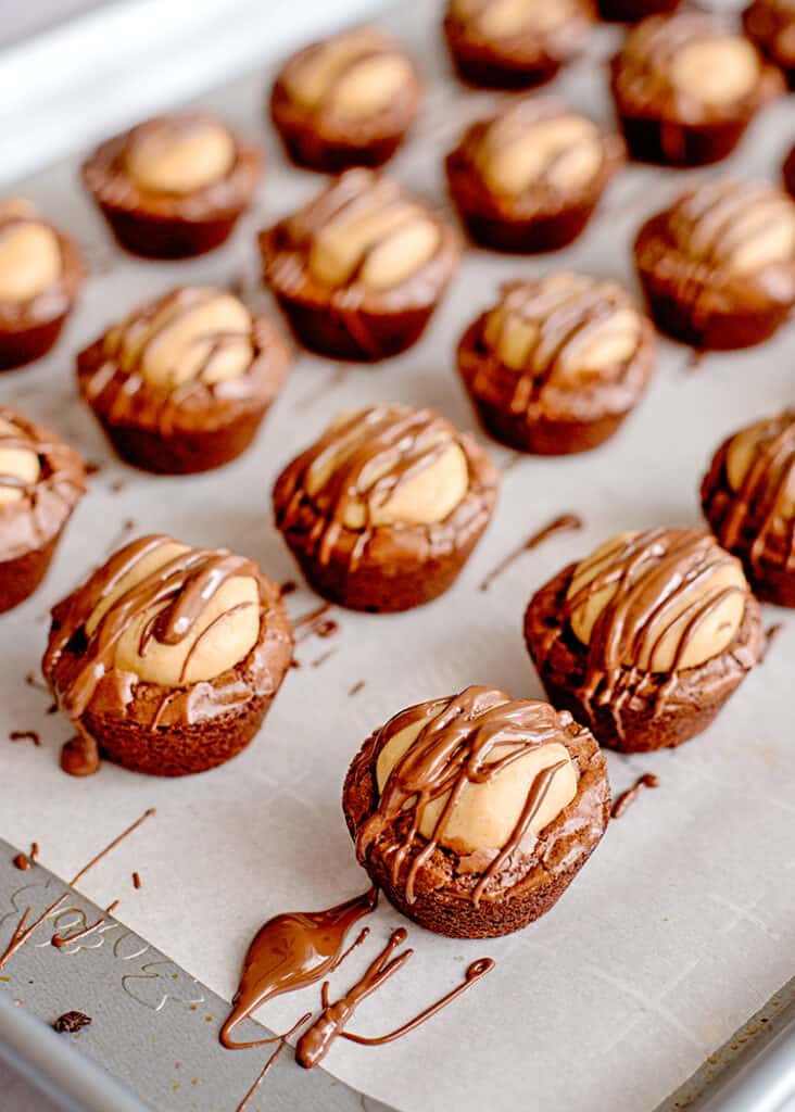Buckeye Brownie Bites - These Buckeye Brownie Bites combine buckeyes and brownies together in a cute little package so you have one incredible chocolate peanut butter dessert. It's a rich fudgy brownie bottom that is topped with a soft peanut butter ball and a chocolate drizzle. They're so easy to make too thanks to a brownie mix shortcut. #cookiedoughandovenmitt #buckeyes #brownies #desserts buckeye brownie bites on a baking sheet with parchment paper