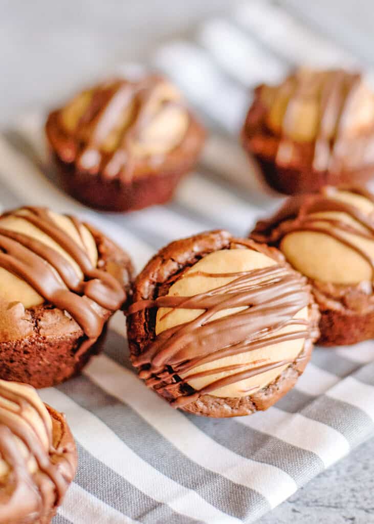 Buckeye Brownie Bites - These Buckeye Brownie Bites combine buckeyes and brownies together in a cute little package so you have one incredible chocolate peanut butter dessert. It's a rich fudgy brownie bottom that is topped with a soft peanut butter ball and a chocolate drizzle. They're so easy to make too thanks to a brownie mix shortcut. #cookiedoughandovenmitt #buckeyes #brownies #desserts front of buckeye brownie bites on a striped linen
