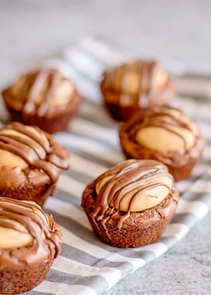 Buckeye Brownie Bites - These Buckeye Brownie Bites combine buckeyes and brownies together in a cute little package so you have one incredible chocolate peanut butter dessert. It's a rich fudgy brownie bottom that is topped with a soft peanut butter ball and a chocolate drizzle. They're so easy to make too thanks to a brownie mix shortcut. #cookiedoughandovenmitt #buckeyes #brownies #desserts side view of buckeye brownie bites on a gray fabric