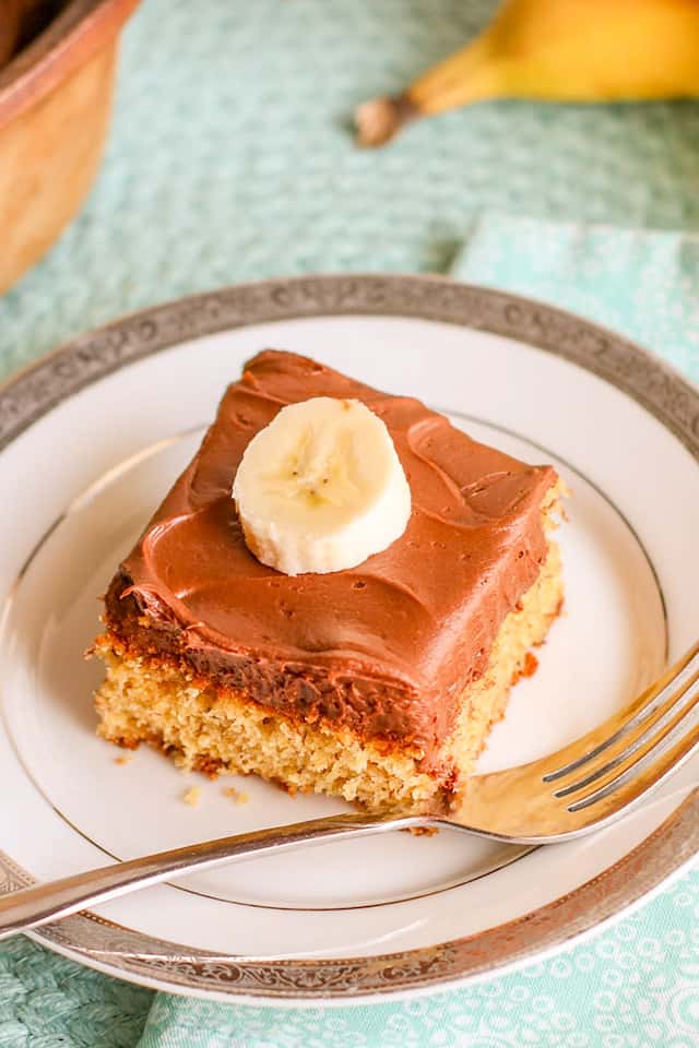 slice of banana cake with chocolate frosting on a white dessert plate with fork