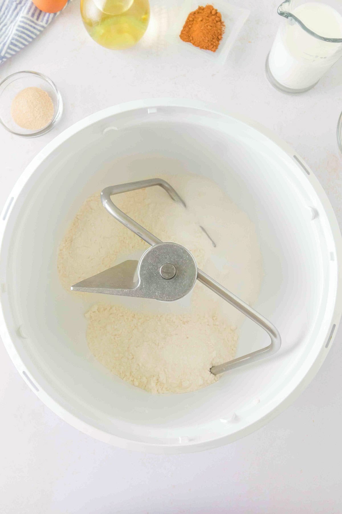 A metal dough hook inside a white stand mixer bowl containing flour for honey buns, surrounded by small containers of ingredients.