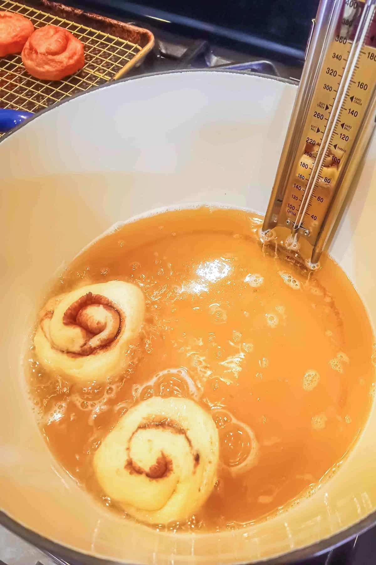 Two raw honey buns frying in a pot of oil with a candy thermometer submerged on the side.