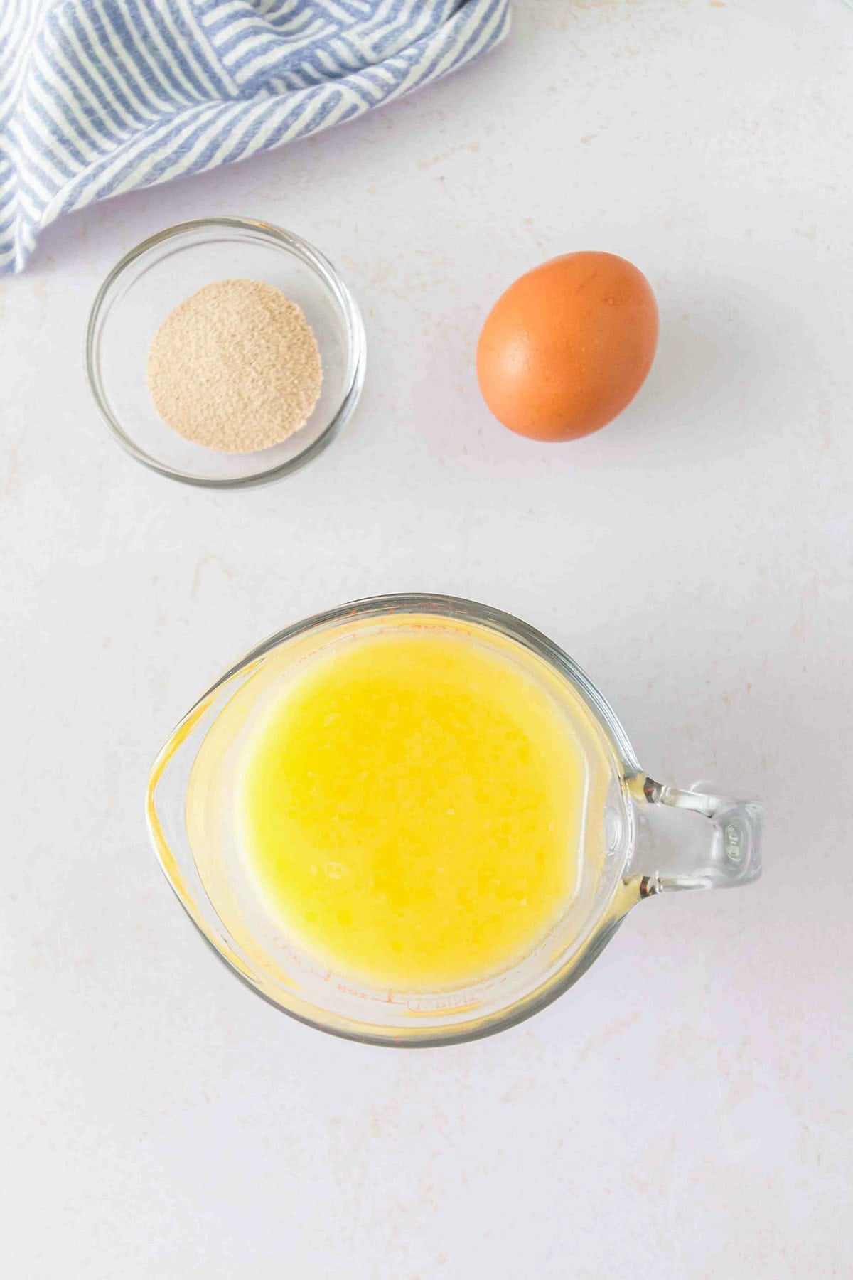 A glass measuring cup containing yellow liquid for honey buns, a small bowl of yeast, and a whole egg on a white surface.