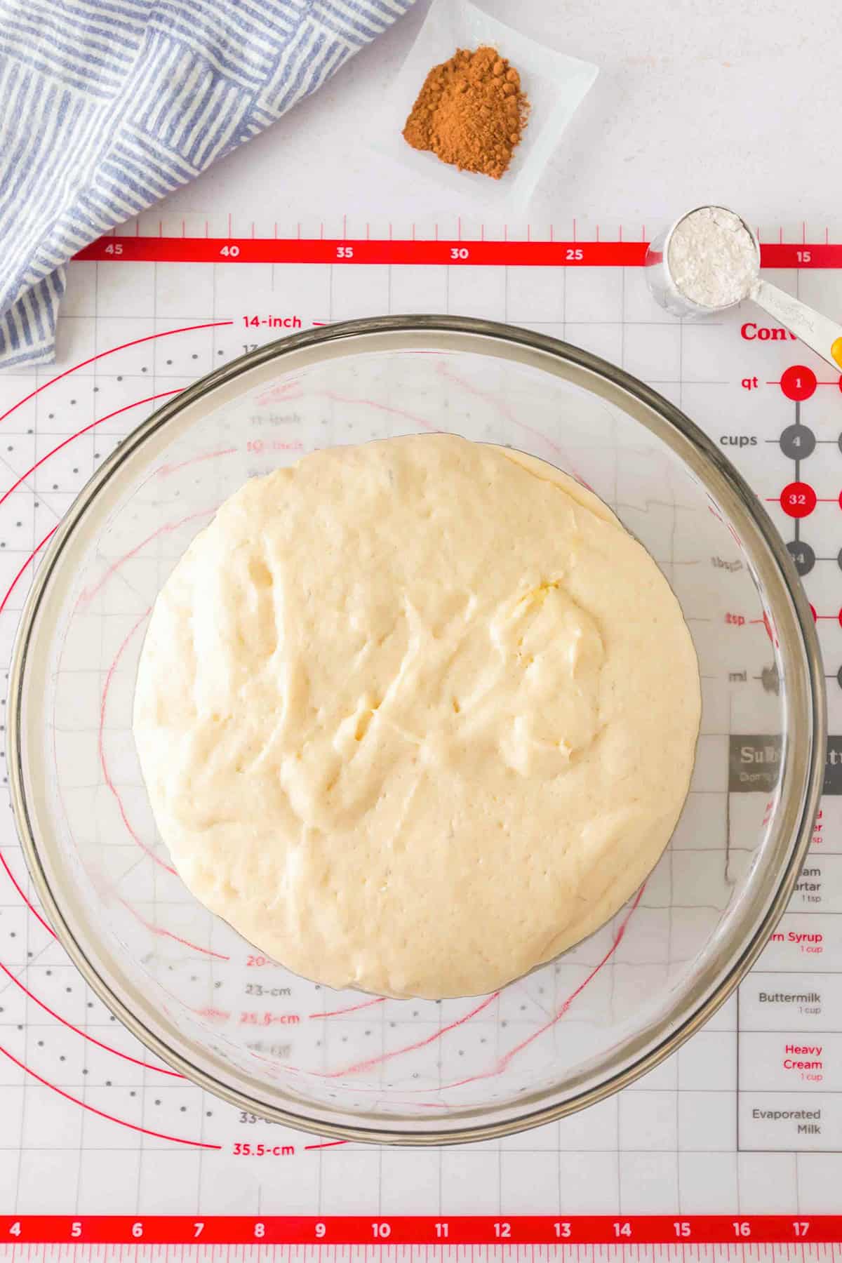 Dough for honey buns rising in a glass mixing bowl on a silicone pastry mat, with a small dish of cinnamon and a measuring cup of flour nearby.