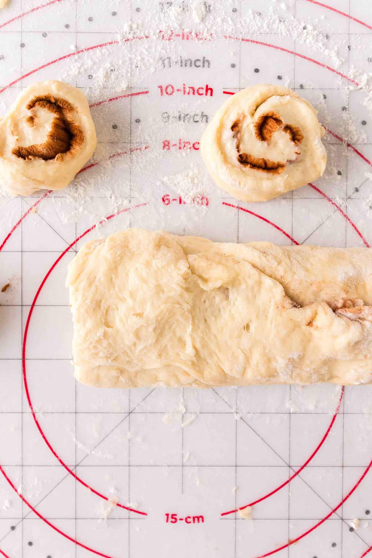 Two honey bun cinnamon rolls and a log of rolled dough on a marked silicone baking mat dusted with flour.