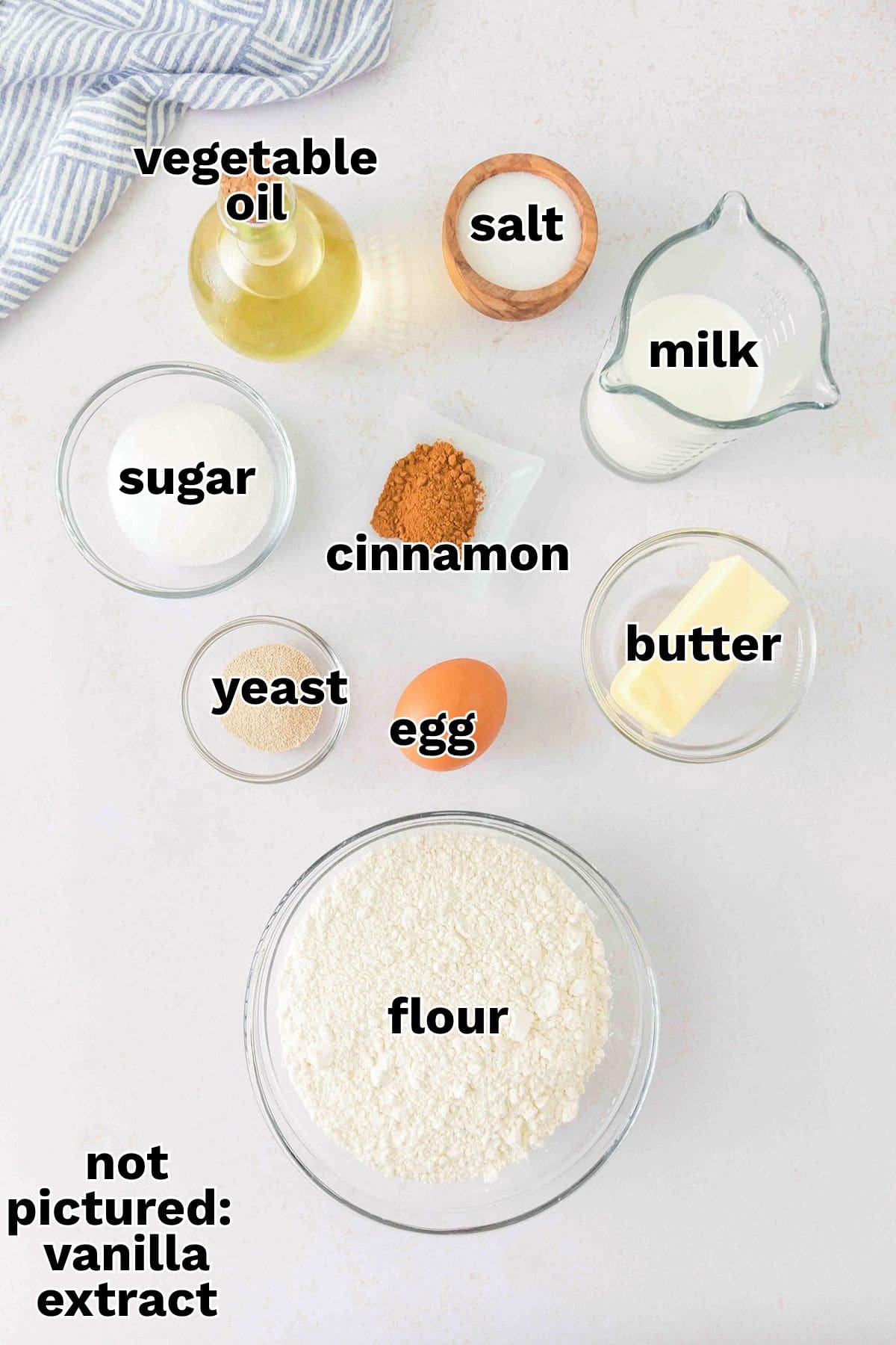 Ingredients for a Honey Buns recipe arranged on a table, including flour, sugar, milk, butter, oil, salt, yeast, cinnamon, and an egg, with a note that vanilla extract is not pictured.