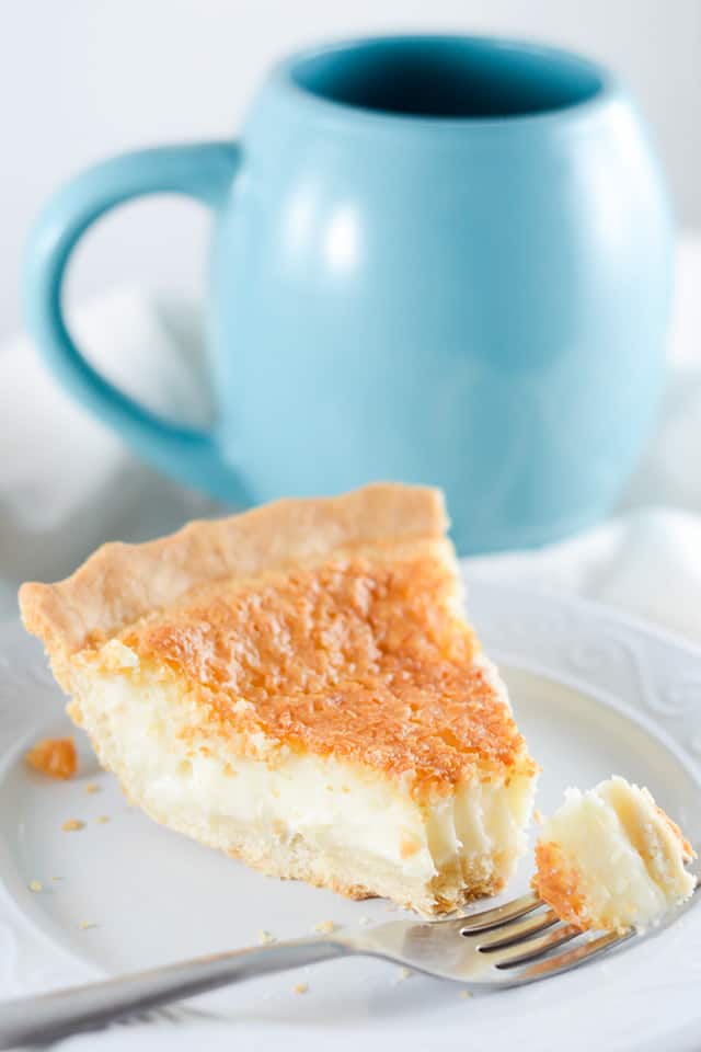 a slice of sugar cream pie with a forkful beside in on a white plate with a blue cup