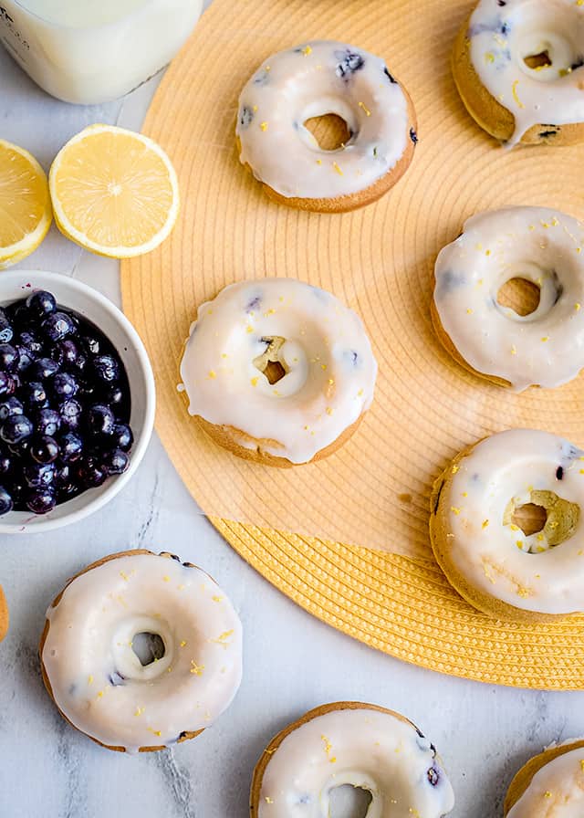 lemon blueberry donuts laid on a marble background with a yellow round place mat with a sliced lemon and blueberries beside the donuts