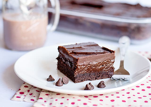 a chocolate bar on a white plate with fork and chocolate chips with chocolate milk and a pan of bars behind it