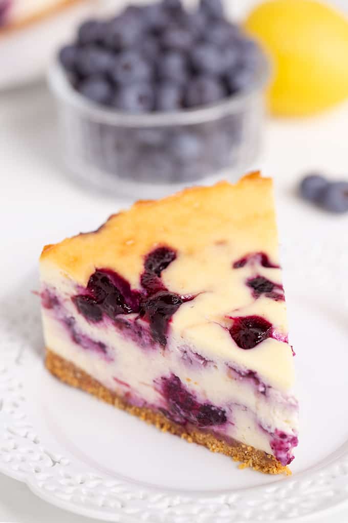 slice of blueberry cheesecake on a white plate with blueberries and lemon behind the plate