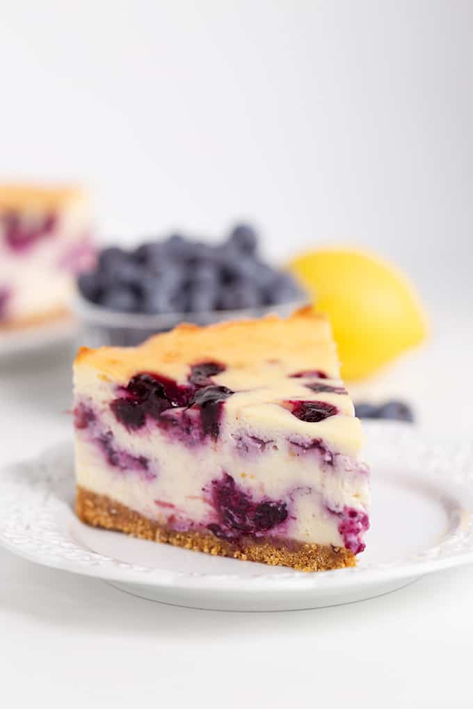 slice of cheesecake on a white plate with a bowl of blueberries and lemon behind the plate on a white surface