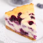 square image of a slice of cheesecake on a white lace plate with a couple of blueberries on the white background behind it