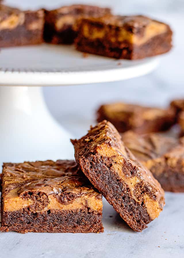 two pumpkin brownies in front of a white cake plate full of more brownies