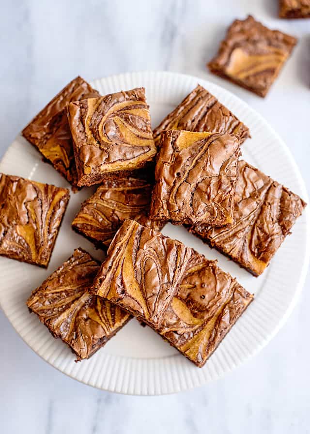 overhead view of pumpkin swirled brownies on a white cake plate and a marble background
