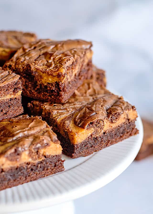 two pumpkin brownies in front of a white cake plate full of more brownies