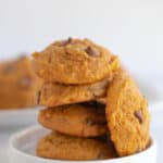 pumpkin chocolate chip cookies stacked on a small white dessert plate with a platter of cookies behind it
