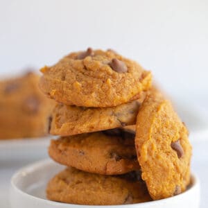pumpkin chocolate chip cookies stacked on a small white dessert plate with a platter of cookies behind it