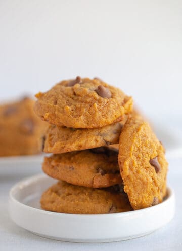 pumpkin chocolate chip cookies stacked on a small white dessert plate with a platter of cookies behind it