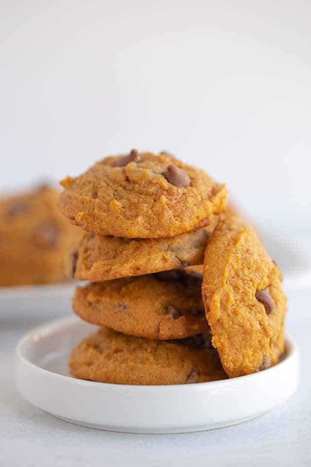 pumpkin chocolate chip cookies stacked on a small white dessert plate with a platter of cookies behind it