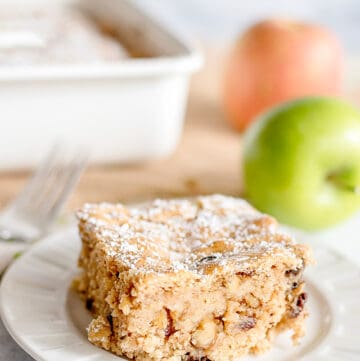 slice of applesauce cake on a white dessert plate with apples and a pan of cake behind it