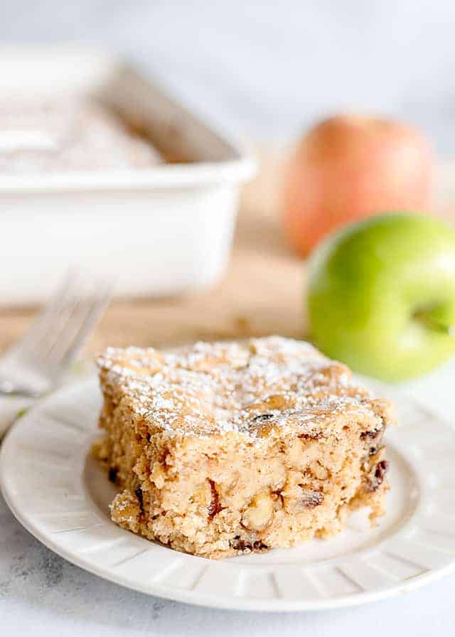 slice of applesauce cake on a white dessert plate with apples and a pan of cake behind it