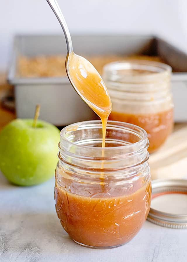 caramel dripping a spoon into a glass jar with an apple behind it