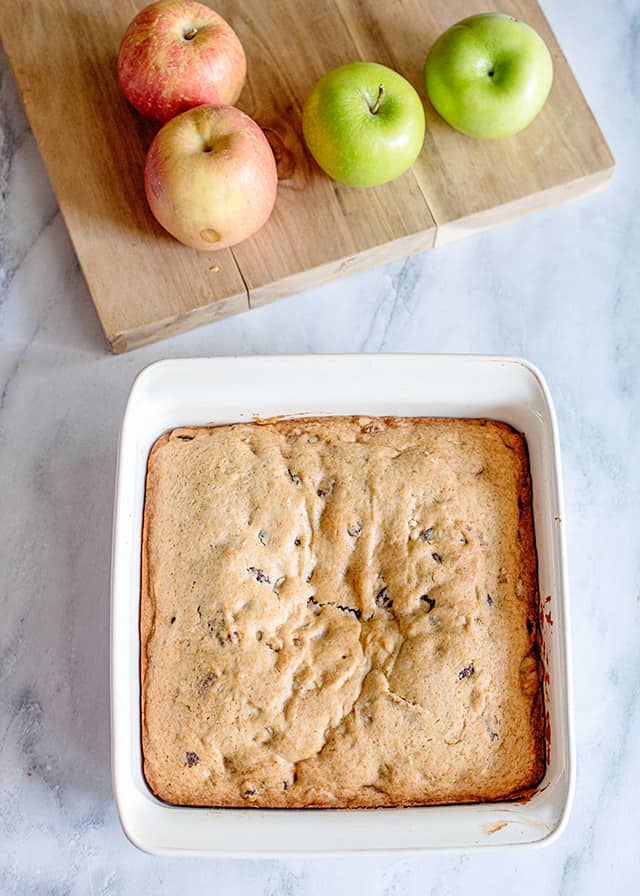 freshly baked applesauce cake in a white pan with a cutting board full of apples