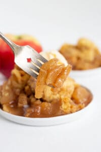 apples on a fork with a plate full of crockpot apple cobbler behind it