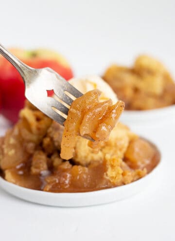 apples on a fork with a plate full of crockpot apple cobbler behind it