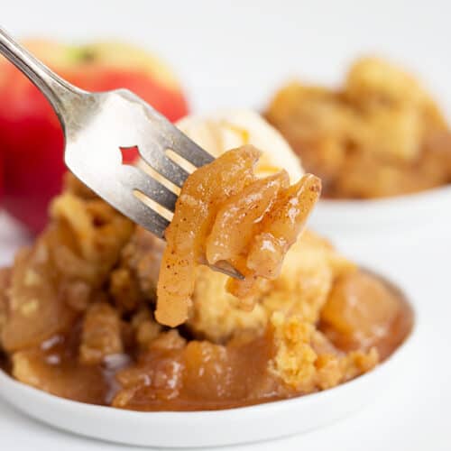 apples on a fork with a plate full of crockpot apple cobbler behind it