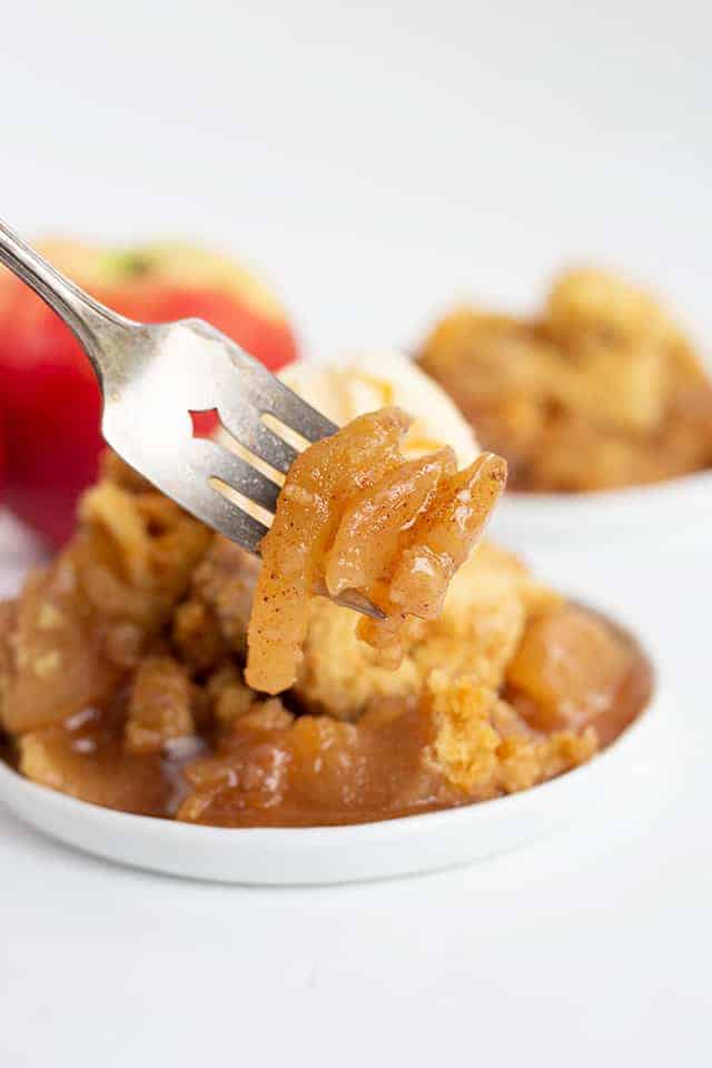 apples on a fork with a plate full of crockpot apple cobbler behind it