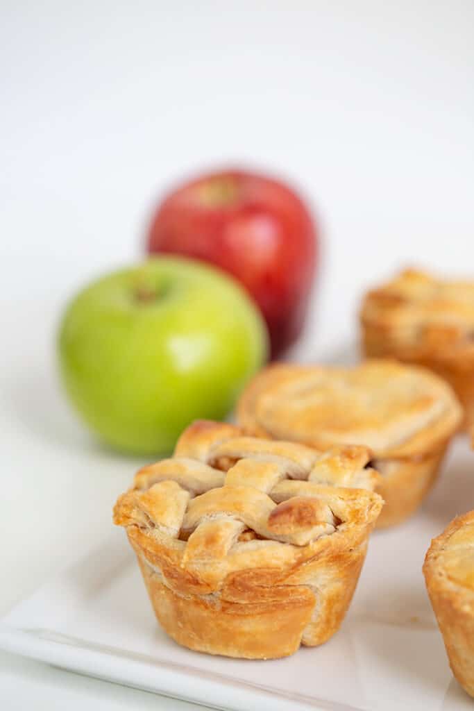 Mini apple pies in a white platter with green and red apples behind them