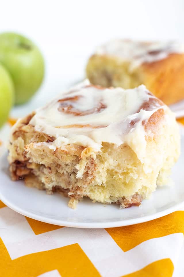 cinnamon roll on a white plate with a yellow and white fabric under it and apples behind the roll