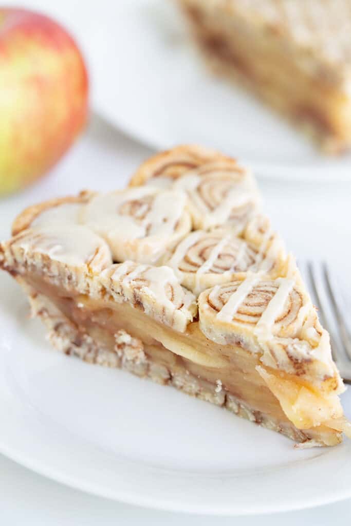 slice of apple pie on a white plate with an apple behind it and a second slice of apple pie