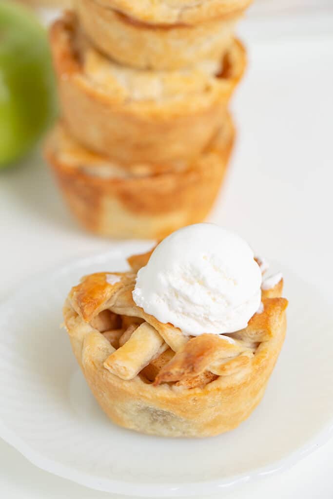 mini apple pie with whipped cream on top on a small white plate with a stack of pies behind it