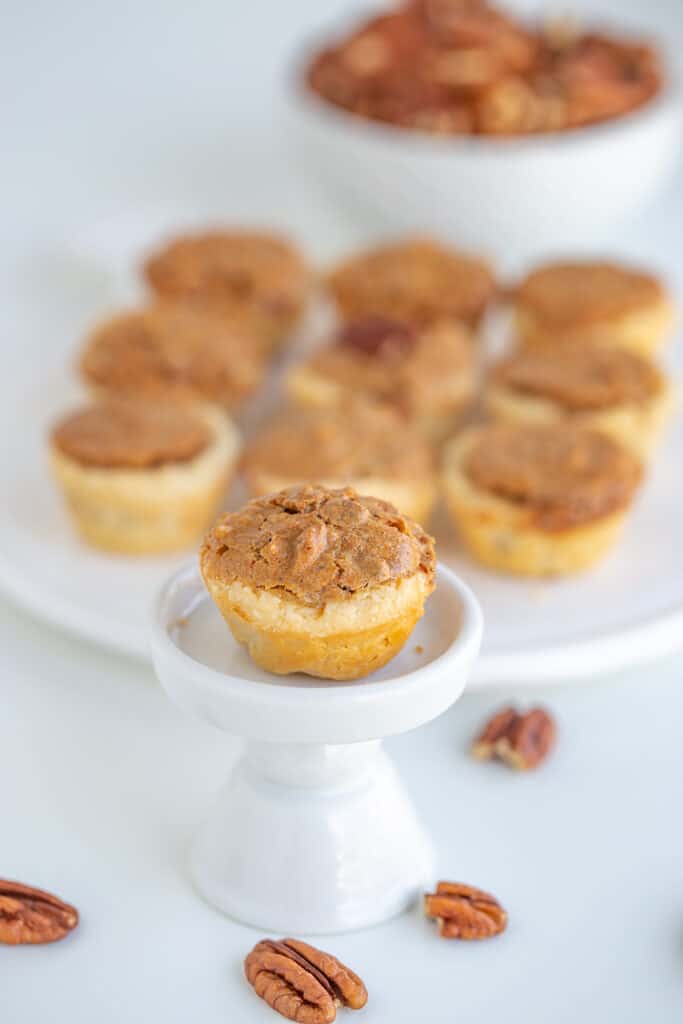 a pecan tassie on a cupcake stand with a platter of tassies behind it and pecans below it