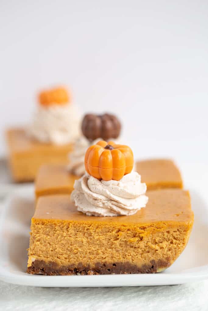 two slices of pumpkin cheesecake bars on white plates with pumpkins on top