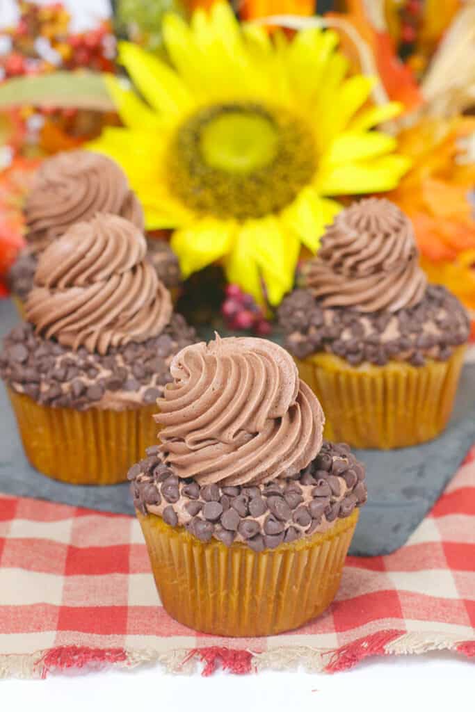 a cupcake with other cupcakes behind it on a dark slate board and flowers behind the cupcakes