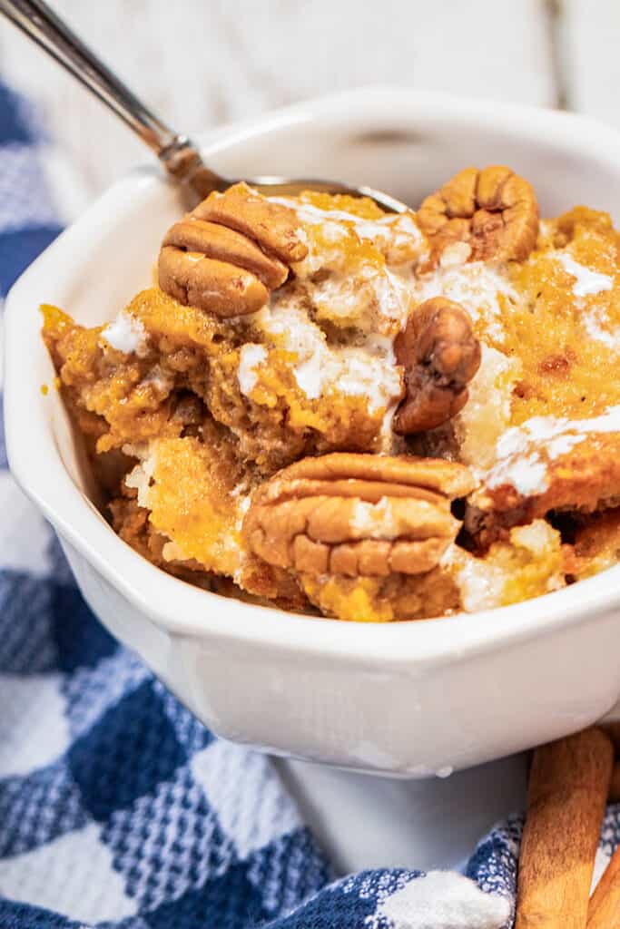 white bowl full of pumpkin pecan cobbler with marshmallow glaze drizzled on top and a blue and white checkered fabric by the bowl