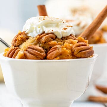 white serving dish filled to the top with pumpkin cobbler, pecans, and whipped cream