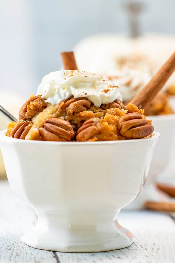 white serving dish filled to the top with pumpkin cobbler, pecans, and whipped cream