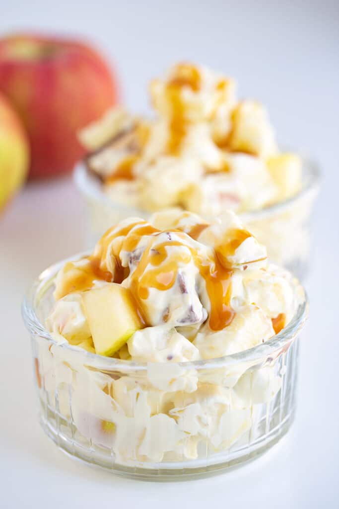 glass bowl full of apple salad on a white surface with apples behind it