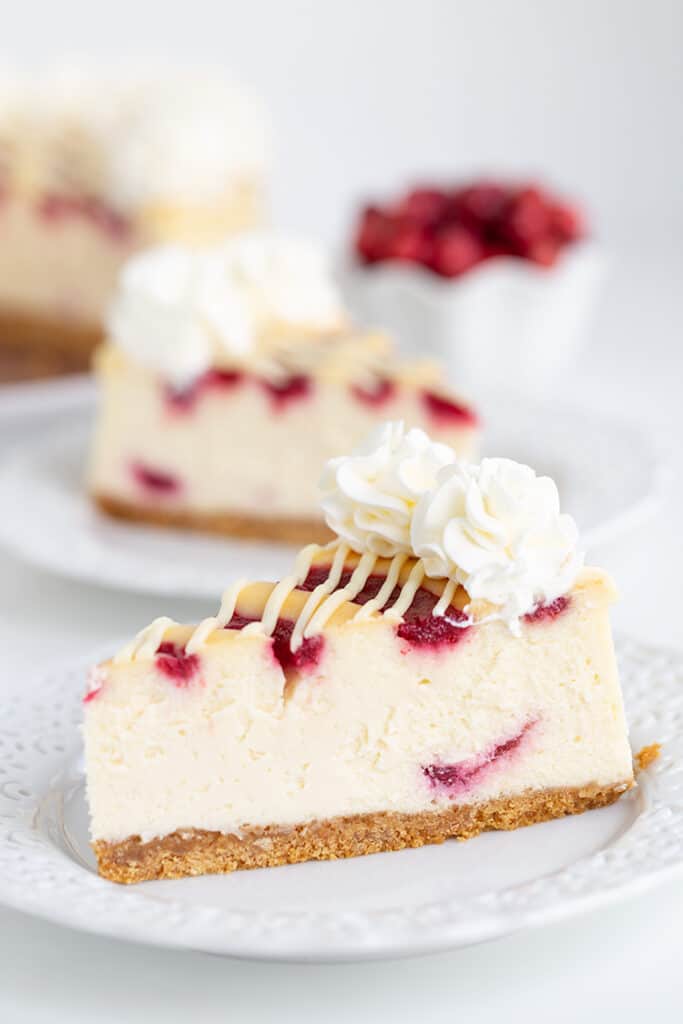 Slice of cheesecake on a white plate with a second slice behind it and cranberries