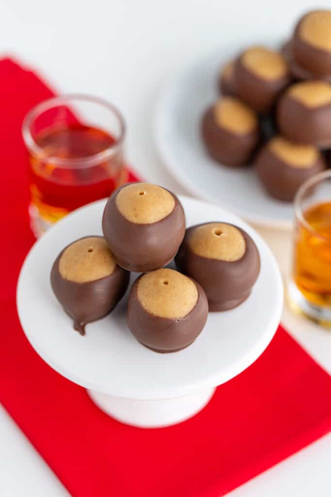 alcoholic peanut butter balls on a white cupcake plate on a red fabric with a shot behind the stand