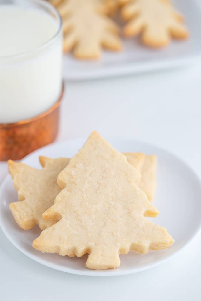 christmas tree cookies on a small white plate with a platter of cookies behind it and a glass of milk