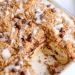 A Chocolate Chip Coffee Cake with a crumb topping and icing drizzle sits in a baking dish, with a square piece removed to reveal its delicious, layered interior.